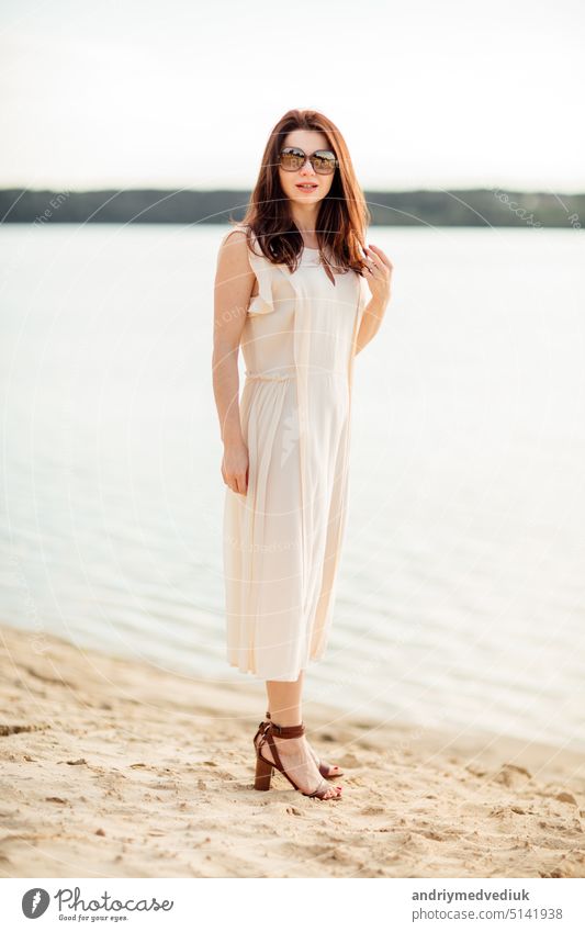 Hübsche junge Frau mit dunklen Haaren in der Nähe am Strand in der Nähe des Sees in Kleid und Sonnenbrille. Konzept der glücklichen Urlaub und Resort Zeit Glück