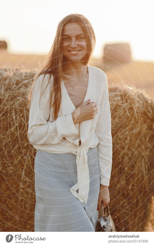 Ganzkörperporträt einer lächelnden schönen Brünetten im Jeansrock und mit einer Strohtasche in der Hand. Frau genießt einen Spaziergang in einem Weizenfeld mit Strohballen an einem sonnigen Sommertag.