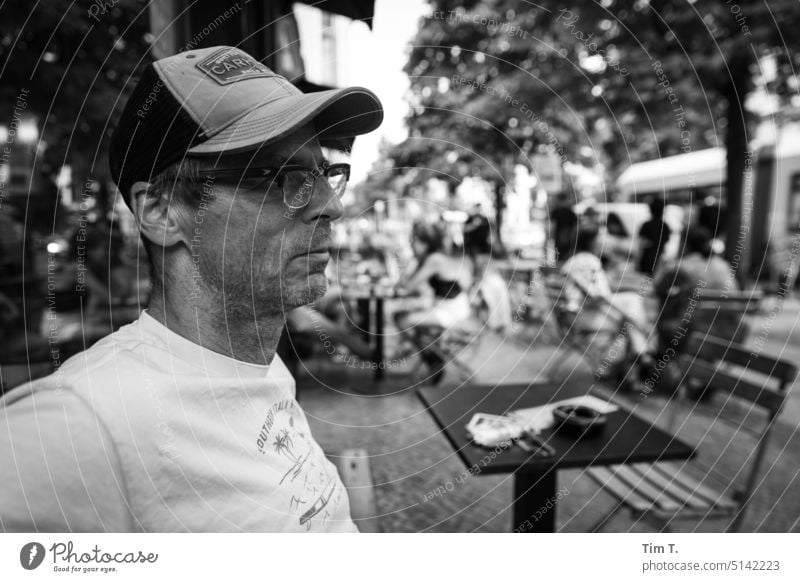 Ein Mann mit Schirmmütze und Brille vor einem Cafe in Berlin Prenzlauer Berg Café kastanienallee Außenaufnahme Stadt Stadtzentrum Altstadt Hauptstadt Tag s/w