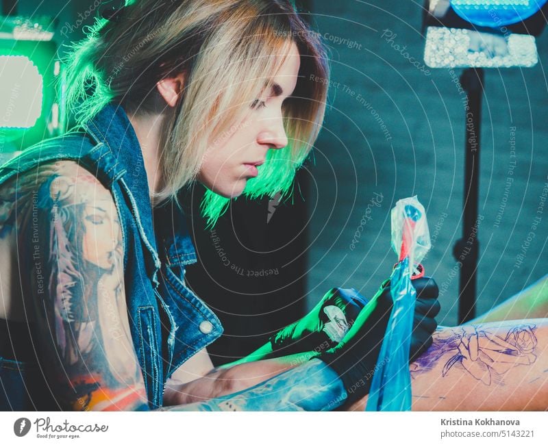 Blick auf professionelle blonde Tattoo Meisterin malt mit schwarzer Tinte, machen Abdeckung. Arbeitet in sterilen Latex-Handschuhen mit handgefertigten Rotor Pistole Maschine im Studio.