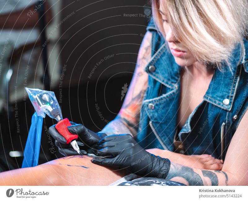Blick auf professionelle blonde Tattoo Meisterin malt mit schwarzer Tinte, machen Abdeckung. Arbeitet in sterilen Latex-Handschuhen mit handgefertigten Rotor Pistole Maschine im Studio.