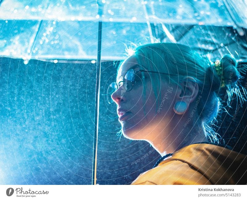 Junges hübsches Mädchen mit blau gefärbtem Haar in gelbem Regenmantel und mit transparentem Regenschirm steht in der Nähe von Brunnen. Nacht Neon-Beleuchtung der Stadt. Porträt von stilvollen Hipster mit Brille.