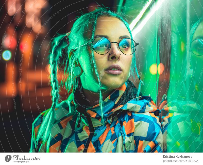Junges hübsches Mädchen mit ungewöhnlicher Frisur in der Nähe glühenden grünen Neonlicht der Stadt in der Nacht. Gefärbte blaue Haare in Zöpfen. Pensive Hipster Teenager mit Brille.