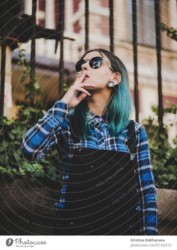 Junges attraktives Punk-Mädchen raucht Zigarette. Hipster mit blauem, ungewöhnlich gefärbtem Haar, Nasenpiercing, violetten Brillengläsern und Ohrentunneln auf einer leeren, alten europäischen Straße.