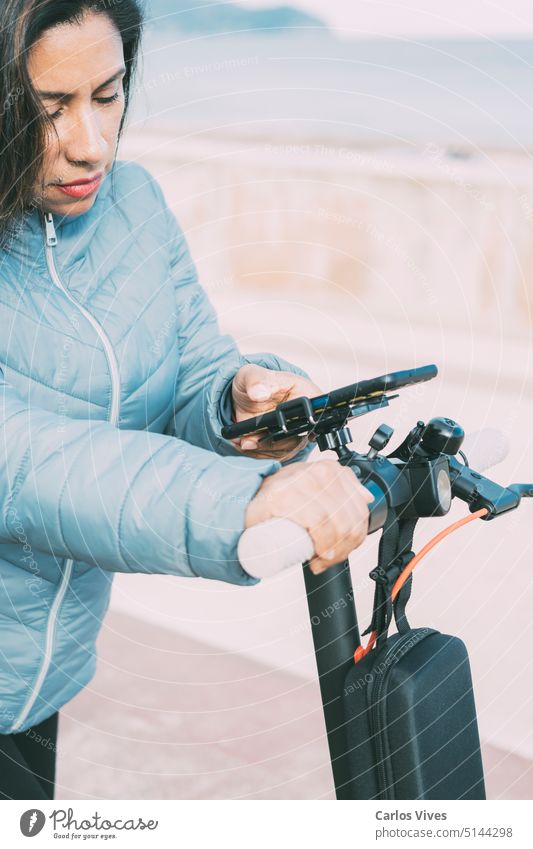 Schöne lateinamerikanische Frau fährt mit ihrem Motorroller durch die Stadt und aktiviert eine Smartphone-App. lässig Großstadt abschließen Tag E-Roller Öko