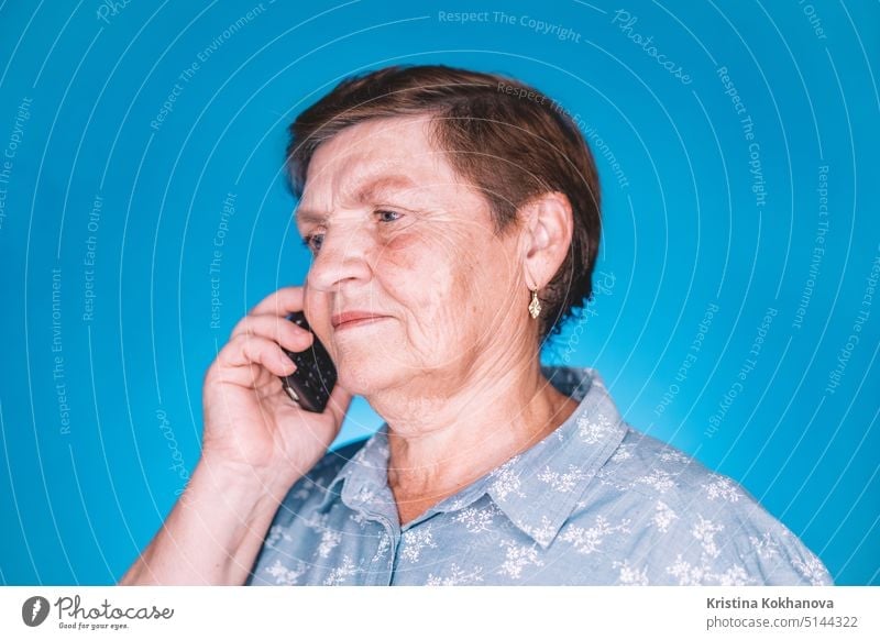 Ältere Rentnerin im Gespräch mit einem mobilen Gerät an einer blauen Studiowand. Großmutter lächelnd. Technologie, alte Menschen Konzept. Handy Mutter 70s