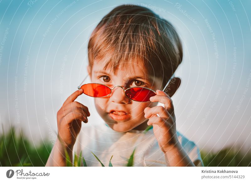 Porträt eines lustigen kleinen Jungen mit roter Sonnenbrille, der in einem frischen grünen Weizenfeld steht. Lovely Kleinkind Kind erforscht Pflanzen, Natur im Frühling. Kindheit, Kinder Mode-Stil-Konzept