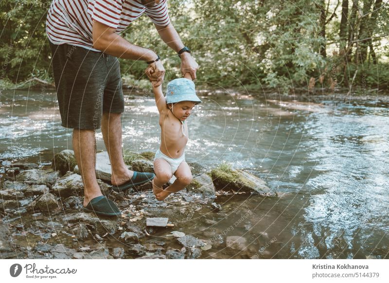 Papa hält sein kleines Baby über den Fluss. Funny Sohn gebunden seine Beine und will nicht im Wasser schwimmen. Familie Vater Menschen Kind Mann Natur Kindheit