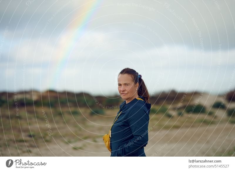 Nachdenklich in die Kamera blickende Frau mittleren Alters mit einem Regenbogen im Hintergrund, Konzept für Zukunftsaussichten nachdenklich Ausdruck Stehen