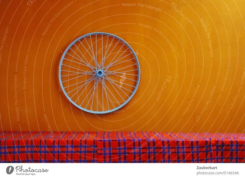 Felge eines Fahrrads auf oranger Wand vor rot-karierter Tischdecke Massai Kunst Speichen Rad rund drehen Decke bunt Verkehr Fahrradfahren