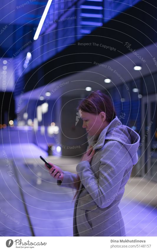 Junge Frau hält Handy, SMS und Nutzung sozialer Medien in blauem Neonlicht in der Stadt bei Nacht, echte Menschen mit Technologie durch das Büro. Moderner Lebensstil in einer großen Stadt