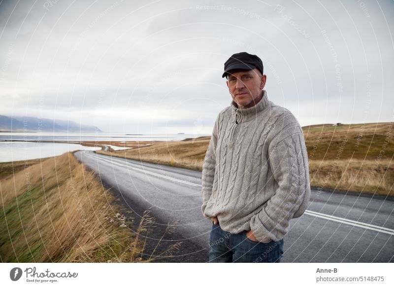 ein Mann steht in einem Strickpullover an einer wirklich einsamen Straße in eine wunderschönen hügeligen Fjordlandschaft  an einem schönen grauen Tag einzeln