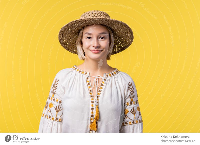 Porträt einer jungen ukrainischen blonden Frau in traditioneller gestickter Bluse auf gelbem Studiohintergrund. Selbstbewusstes sonniges Outfit, Strohhut.
