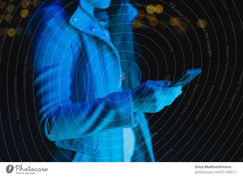 Unerkennbare Frau, die ein Handy hält, eine SMS schreibt und soziale Medien in blauem Neonlicht in einer nächtlichen Stadt nutzt. Echte Menschen nutzen Technologie. Frau steht draußen mit Smartphone in den Händen.