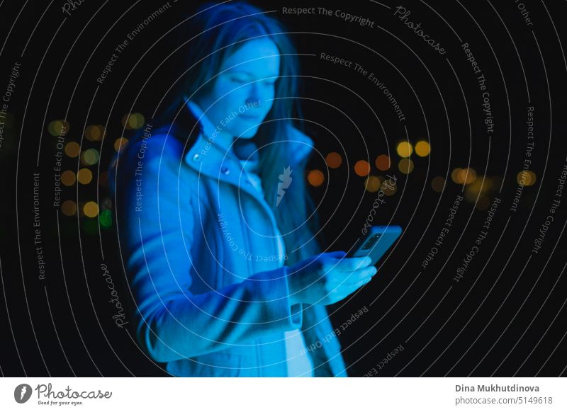Frau hält Handy, SMS und Nutzung sozialer Medien in blauem Neonlicht in der Stadt bei Nacht. Echte Menschen nutzen Technologie. Frau steht draußen mit Smartphone in ihren Händen.