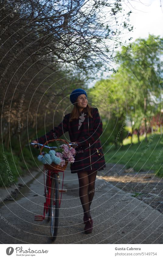 Ein lächelndes Mädchen geht mit ihrem roten Fahrrad und Blumen einen Weg mit Bäumen entlang. Park grün Natur genießend Lifestyle Frau jung Lächeln im Freien