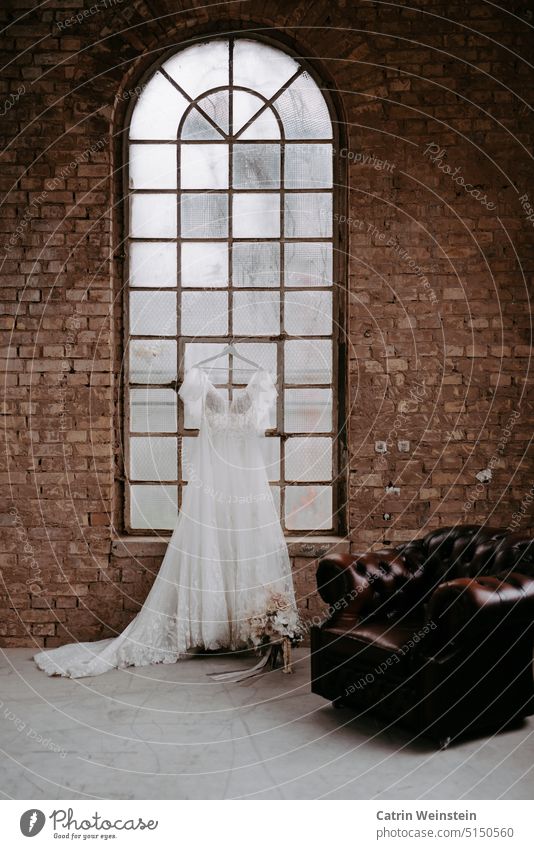 Ein Brautkleid hängt an einem Bügel an einem Rundbogenfenster. Davor steht ein Brautstrauß und ein alter Ledersessel. Backsteinwand weiß rot Sessel rosa Wand