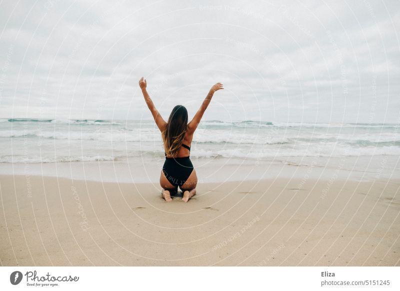 Frau im schwarzen Badeanzug kniet mit ausgestreckten Armen und Rücken zur Kamera am Strand und blickt aufs Meer ausgestreckte Arme Freude Freiheit Urlaub