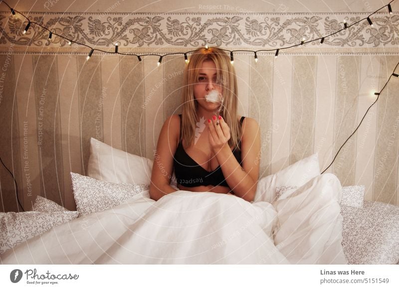 Ein schönes blondes Mädchen in schwarzen Dessous raucht eine Zigarette. Sie sitzt in ihrem Bett mit weißen Laken und Kissen um sie herum. Sie flirtet mit einer Kamera. Zeigt ihr wunderschönes Aussehen und ihre sexy Kurven.
