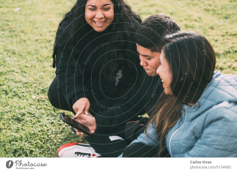 Gruppe von Latinos, die lachend mit einem Smartphone auf dem Boden in einem Park sitzen Zugänglichkeit Argentinien Tasche Bonden Rundfunksendung sorgenfrei