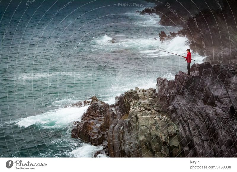 Junge mit rotem Pullover und Angel auf Felsenküste am Meer Angeln Angelrute Junger Mann düster Wellen Ozean Atlantik Atlantikküste Angler Felswand Irland Wasser