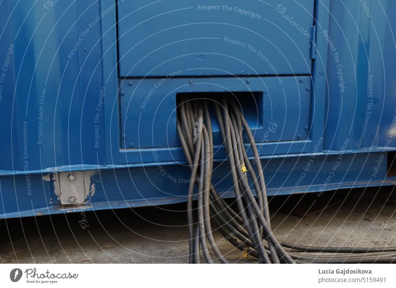 Nahaufnahme eines Bündels schwarzer Kabel, die aus einem blauen Stromaggregat oder einem tragbaren Generator für die Stromversorgung der Kühltransporter an Deck eines beladenen Containerschiffs, das den Ozean durchquert, herauskommen.