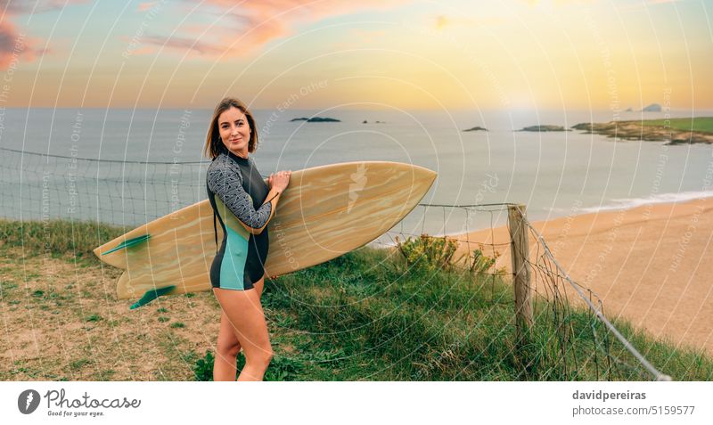 Surferin mit Neoprenanzug posiert mit Surfbrett und schaut in die Kamera in der Nähe des Strandes Frau jung schauende Kamera posierend nah Sonnenuntergang