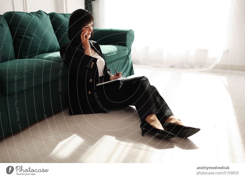 Geschäftsfrau spricht mit einem Smartphone. Tired brunette Frau sitzt in der Nähe von grünen Sofa in einem Geschäft schwarzen Anzug und Schuhe mit hohen Absätzen mit Notebook. Grüne Zimmerpflanze im Interieur. Arbeit von zu Hause aus.