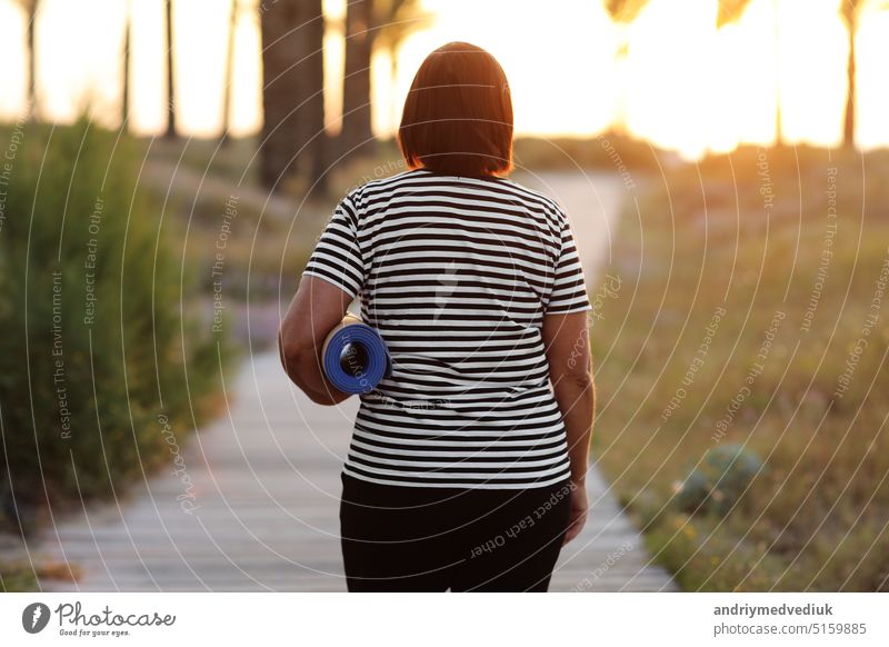 Rückenansicht von aktiven übergewichtigen Frau mit Yoga-Matte zu Fuß zum Strand zu üben Yoga, üben im Freien auf Sonnenaufgang am Morgen. Konzept der gesunden Lebensweise, Wohlbefinden und Fitness in der Natur.