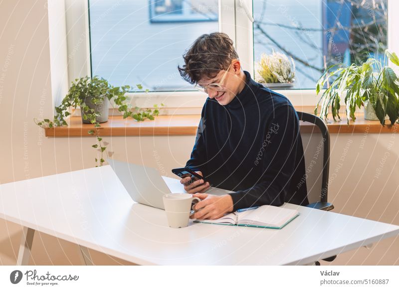 Ein zielstrebiger, braunhaariger Unternehmer mit einem Lächeln im Gesicht antwortet seinen Kunden in seinem Büro mit viel Tageslicht auf sein Mobiltelefon. Von zu Hause aus arbeiten