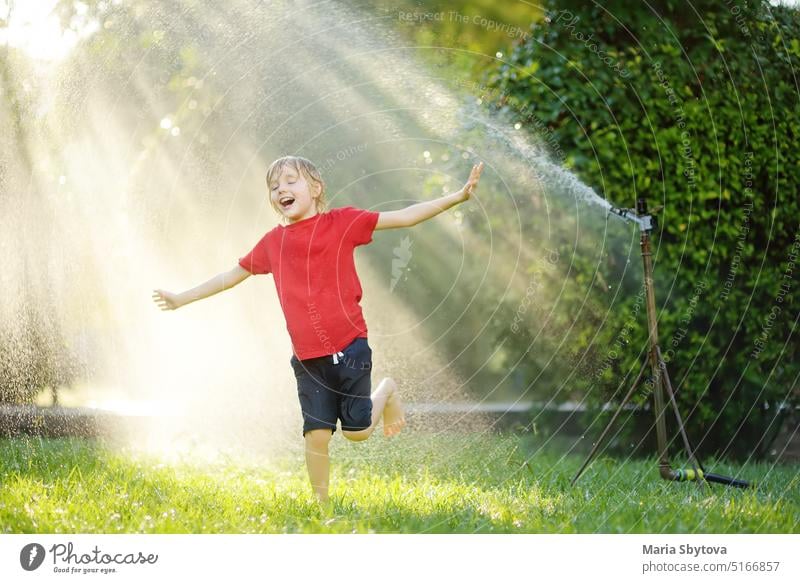 Lustiger kleiner Junge spielt mit Garten Sprinkler in sonnigen Hinterhof. Grundschulkind lacht, springt und hat Spaß mit Sprühwasser. Sommer im Freien Aktivität für Kinder.