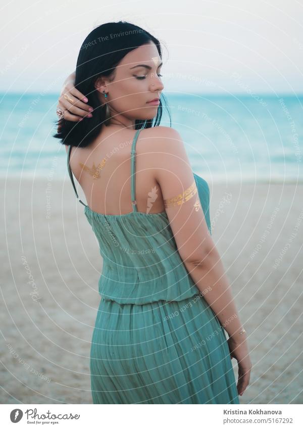 Outdoor-Mode Porträt der schönen Brünette Dame am Strand mit Flash-Tattoos auf Händen, Rücken und Stirn. Zigeuner Boho-Stil. Frau im langen grünen Kleid schaut in die Kamera.