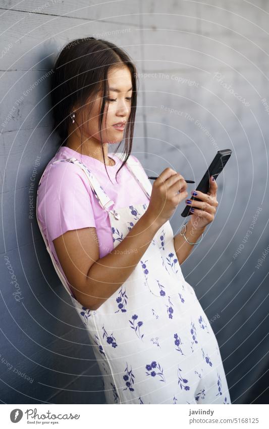 Asiatische Frau lehnt sich an die Wand und benutzt ein Mobiltelefon Smartphone benutzend Griffel fettarm Stil Nachrichten Straße soziale Netzwerke jung ethnisch