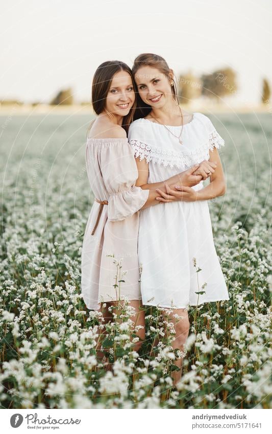 Porträt der schönen jungen zwei Frauen Schwestern in weißen Kleidern mit langen Haaren in einem Feld mit weißen Wildblumen im Sommer. Rustikaler Stil, Dorf. Zärtlichkeit, Umarmungen, Unterstützung.