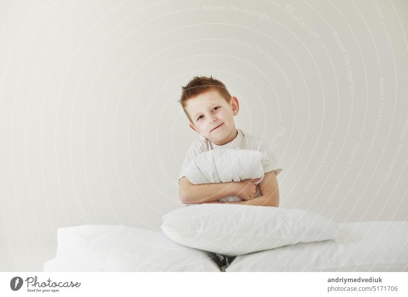 Porträt eines leger gekleideten kleinen Jungen, der sich an ein weißes Kissen schmiegt und sich nach einem langen, anstrengenden Tag müde fühlt, mit einem fröhlichen Lächeln, der sich ausruht