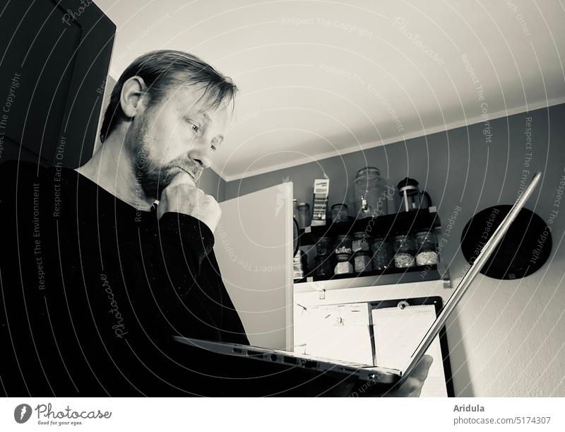 Nachdenklicher Mann arbeitet stehend in der Küche mit Laptop in der Hand in s/w Arbeit nachdenklich Homeoffice Improvisation arbeiten Notebook online Computer