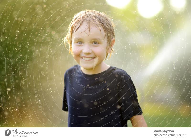 Close-up-Porträt von lustigen kleinen Jungen spielen mit Garten Sprinkler in sonnigen Hinterhof. Grundschulkind lacht, springt und hat Spaß mit Wasser. Sommer im Freien Aktivität für Kinder.