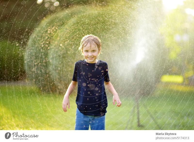 Lustiger kleiner Junge spielt mit Garten Sprinkler in sonnigen Hinterhof. Grundschulkind lacht, springt und hat Spaß mit Sprühwasser. Sommer im Freien Aktivität für Kinder.