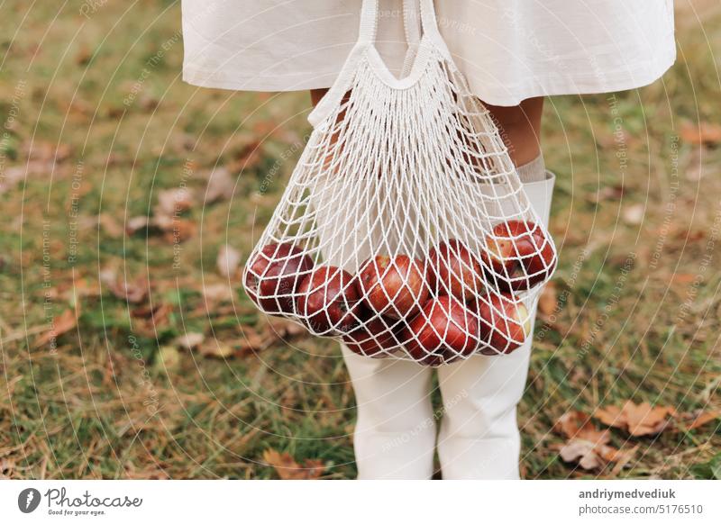 Schnurbeutel mit roten Äpfeln. Blick auf weibliche Beine in weißen Stiefeln, die mit einer Ökotasche in den Händen durch den Herbstgarten laufen. Null-Abfall-Konzept. Kein Leben in Plastik. Gesunder Fitness-Lebensstil. Recycling von Abfall