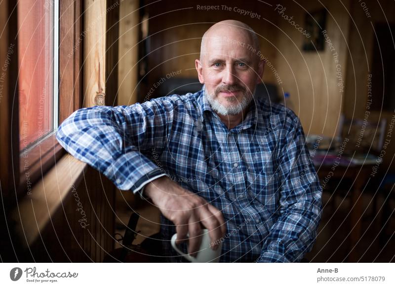 ein Mann mit Bart und Karohemd sitzt zufrieden blickend in einem einfachen Holzhaus am Fenster mit der Kaffeetasse in der Hand. freundlich direkter Blick