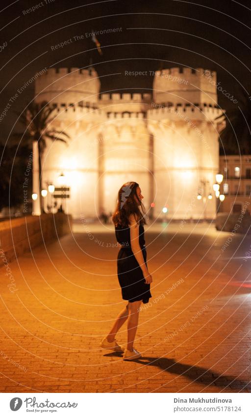 Glückliche Frau zu Fuß in der Nacht in der europäischen Stadt vor einem beleuchteten Schloss, lächelnd. Glückliche Frau zu Fuß in der historischen Mitte der Stadt mit Lichtern. Glücklich lächelnd, auf dem Weg zur Party. Junge und schöne Touristin in Spanien.