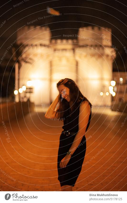 Glückliche Frau zu Fuß in der Nacht in der europäischen Stadt vor einem beleuchteten Schloss, lächelnd. Glückliche Frau zu Fuß in der historischen Mitte der Stadt mit Lichtern. Glücklich lächelnd, auf dem Weg zur Party. Junge und schöne Touristin in Spanien.
