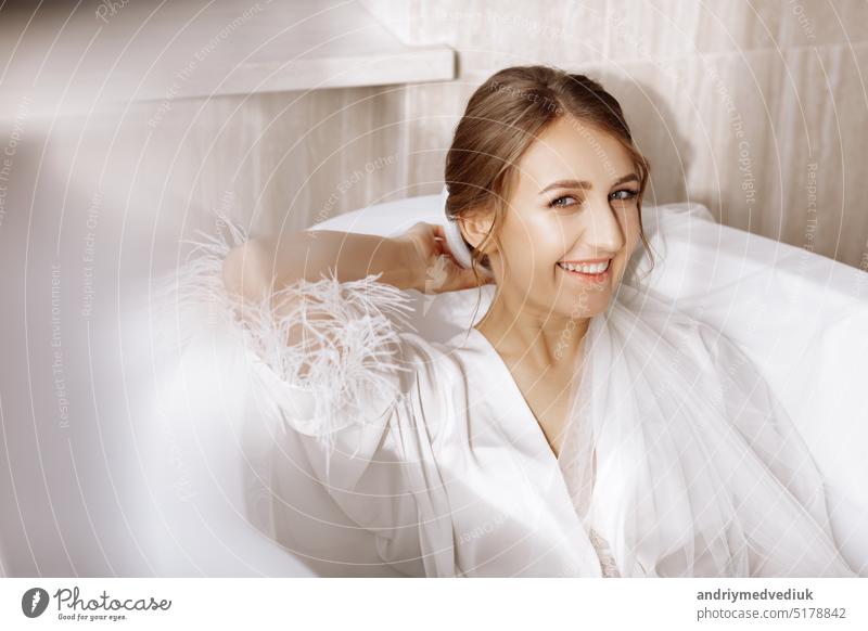 Morgen der Braut. Glückliche schöne junge Frau trägt in einem weißen langen Schleier, Bademantel und Unterwäsche sitzen lachend und mit Spaß in der Badewanne auf einem weißen Hintergrund, entspannen.