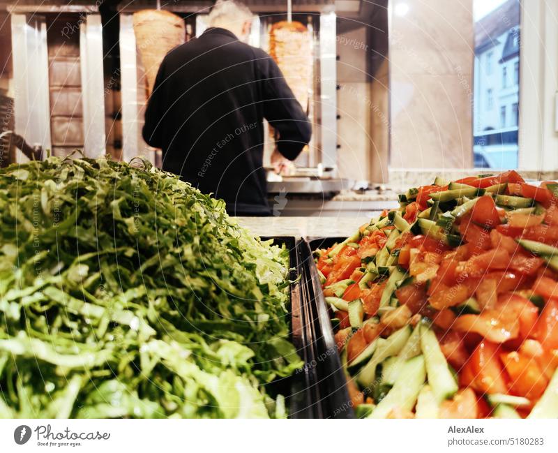 Grüner Salat und Tomaten-Gurkensalat vor zwei Dönerspießes in einem Imbiss, in dem ein Mitarbeiter am Dönerspieß steht Imbiß Essen Fleisch Kalbsfleisch