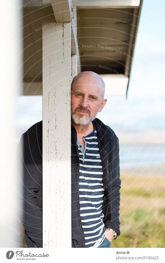 ein Mann mit Bart und Ringelshirt lehnt lässig hinter einem Balken auf der Veranda eines Hauses an einem Gewässer. Er blickt direkt in die Kamera. Portrait