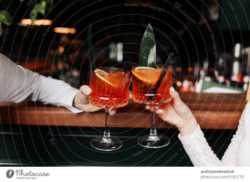 Hände von Frau und Mann klirren, Prost mit Gläsern von Spritz Cocktail. Paar feiert Hochzeit, Jahrestag mit Aperol Spritz Cocktails, mit Orange und Grüns Erfrischendes alkoholisches Getränk