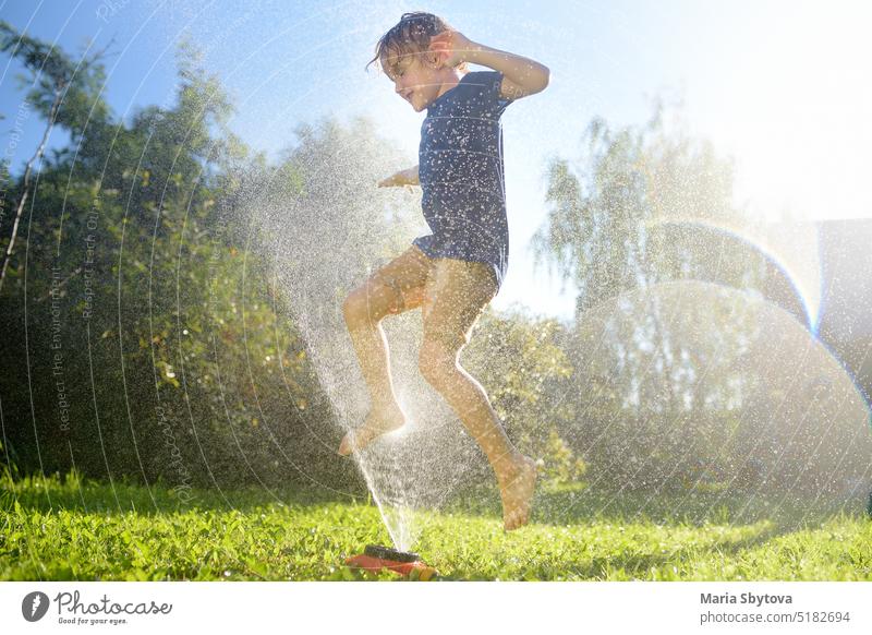 Lustiger kleiner Junge spielt mit Garten Sprinkler in sonnigen Hinterhof. Vorschulkind lacht, springt und hat Spaß mit Sprühwasser. Sommerferien für Kinder auf dem Dorf