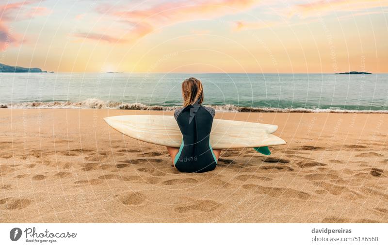 Surferin mit Neoprenanzug und Surfbrett auf dem Sand sitzend und aufs Meer blickend unkenntlich Frau jung Sitzen Blick MEER Rücken Strand Menschen 20s im Freien
