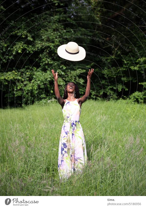 lachende Frau mit fliegendem Strohhut in einer Wiese feminin Park Kleid Hut langhaarig dunkelhaarig stehen Freude Glück schön Fröhlichkeit frei Lebensfreude