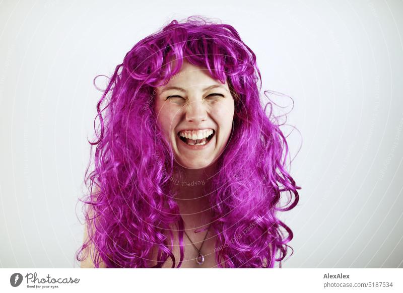 Nahes Portrait einer jungen, blonden Frau, die  vor einem weißen Hintergrund lacht und violette Haare hat frisch Blick in die Kamera Erwartung drinnen